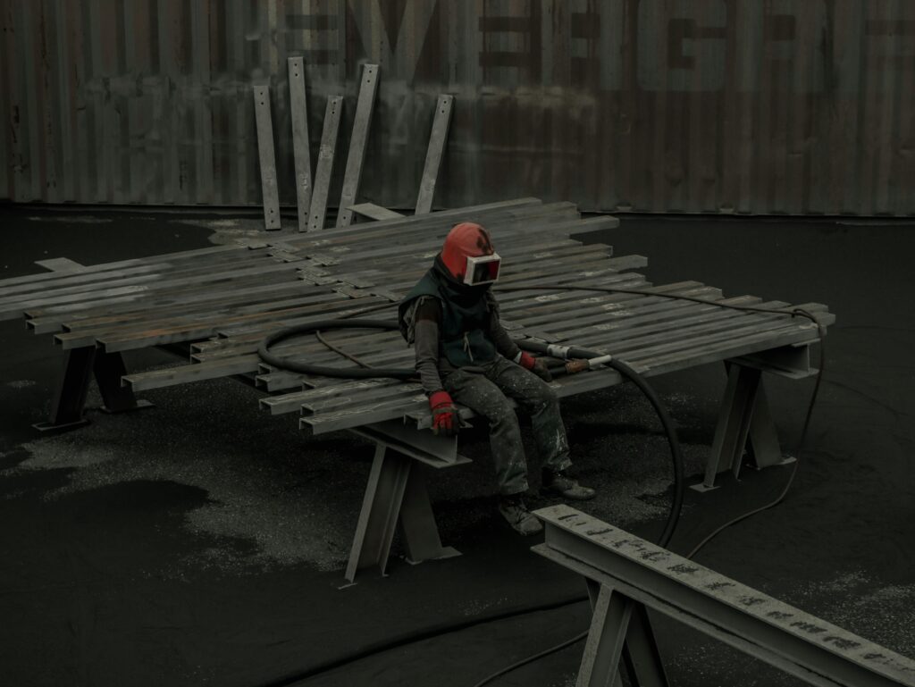 Worker in protective gear resting on metal beams in an industrial setting.